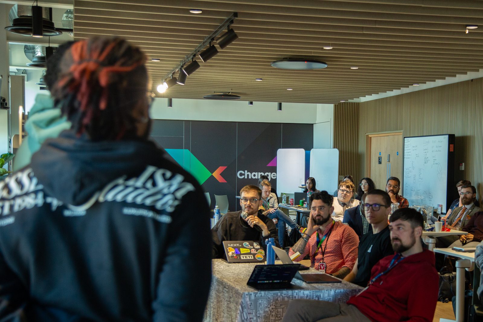 View from behind a presenter towards the hackathon audience seated at tables with laptops, in a room with a 'Change' wall graphic