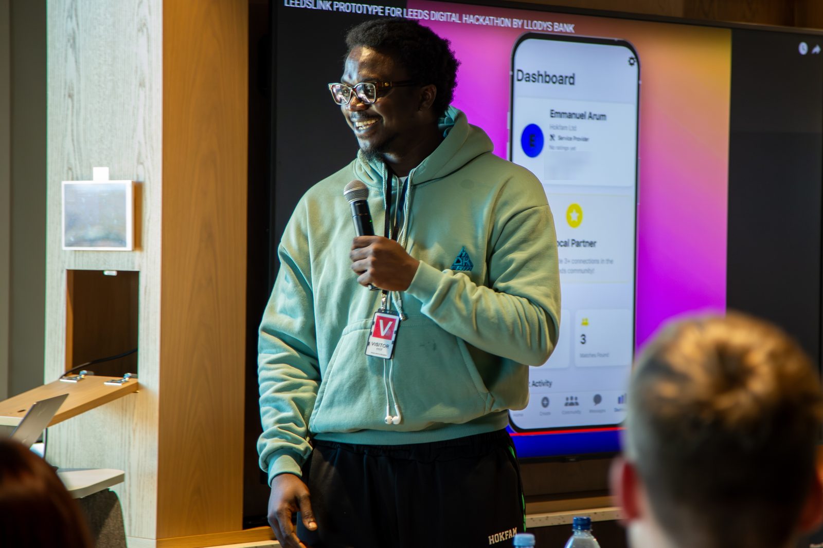 Ikechukwu smiling with a microphone beside a screen showing the LeedsLink dashboard for a Hokfam Ltd service provider profile