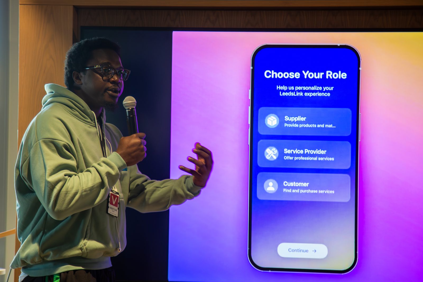 Ikechukwu holding a microphone and gesturing towards a large screen showing the LeedsLink app's 'Choose Your Role' screen, with Supplier, Service Provider and Customer options